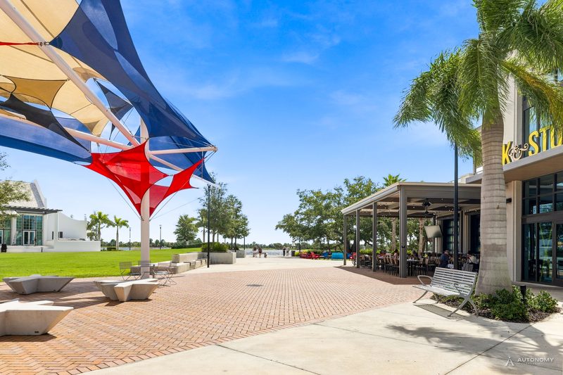 Red, white and blue shade sails over the brick plaza in Downtown Wellen, Wellen Park, with the lawn and Grand Lake beyond